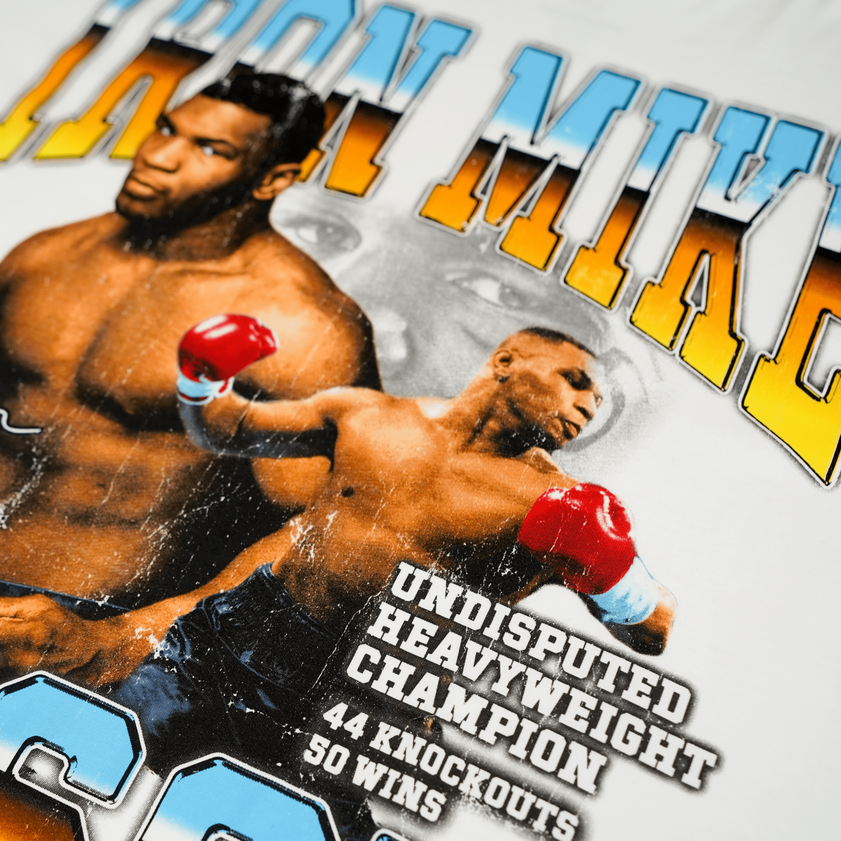 White T-shirt with three images if Mike. One is mid-fight, the other is posing for a photo, and the third is of his eyes in the background. Text above and below the image says Iron Mike Tyson, Undisputed Heavyweight Champion. 44 Knockouts, 50 wins. His signature is on the left.
