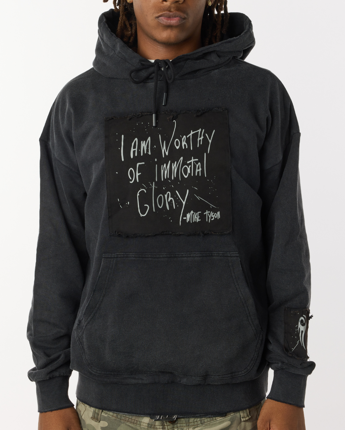 Black distressed hoodie with front pockets. A large black patch on the chest highlights Mike Tyson's handwriting that states "I am worthy of Immortal Glory." A small patch on the left arm displays the Tyson "hook" logo. A medium patch on the back bottom right displays the full Tyson logo.