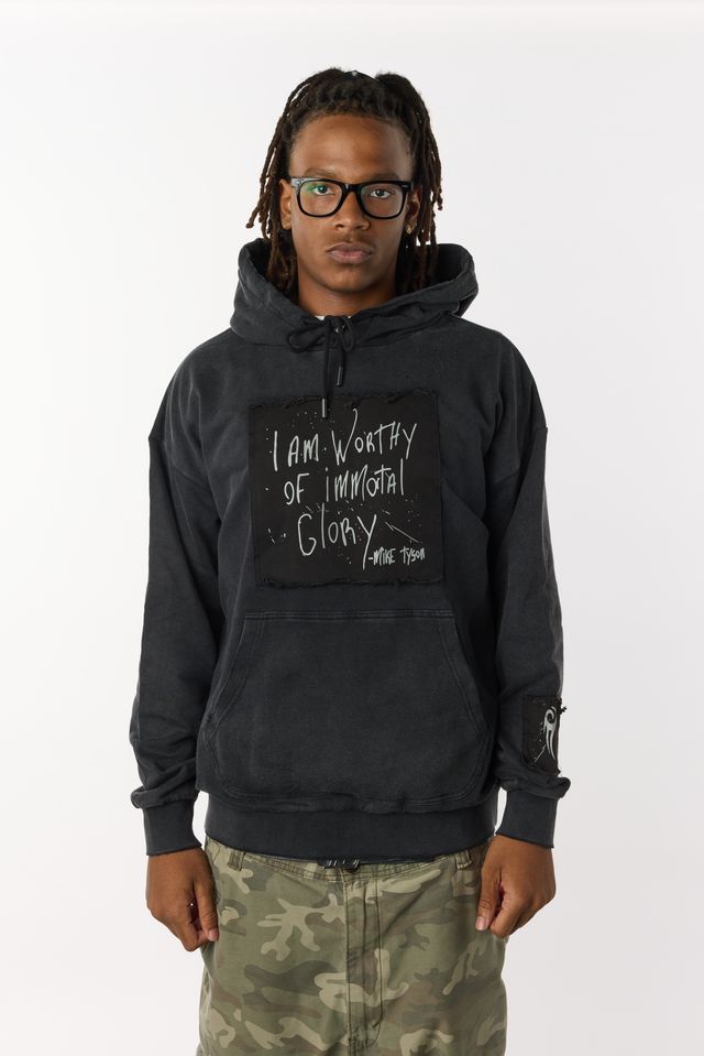 Black distressed hoodie with front pockets. A large black patch on the chest highlights Mike Tyson's handwriting that states "I am worthy of Immortal Glory." A small patch on the left arm displays the Tyson "hook" logo. A medium patch on the back bottom right displays the full Tyson logo.