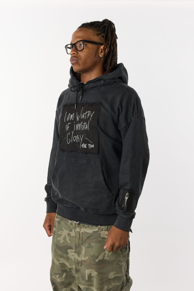 Black distressed hoodie with front pockets. A large black patch on the chest highlights Mike Tyson's handwriting that states "I am worthy of Immortal Glory." A small patch on the left arm displays the Tyson "hook" logo. A medium patch on the back bottom right displays the full Tyson logo.