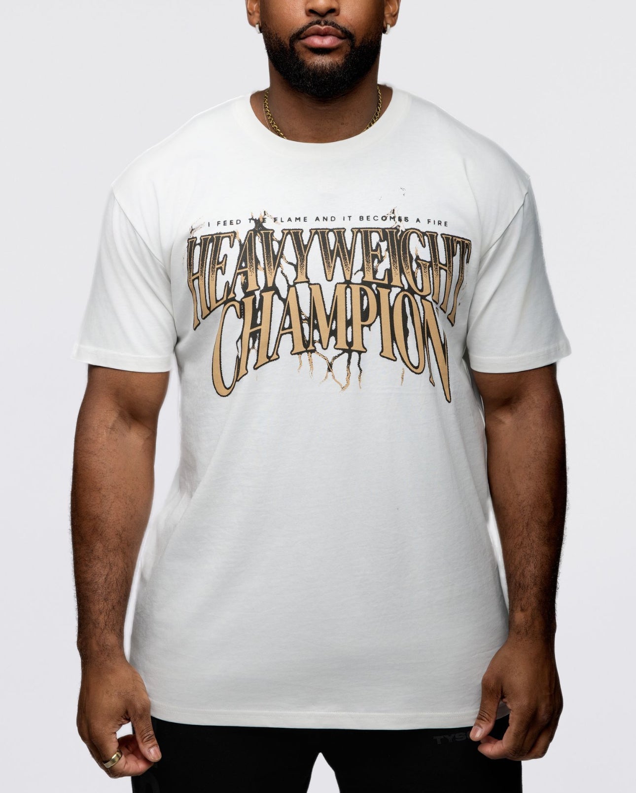 The front of this white shirt features the text "Heavyweight Champion" in a bronze ombre. Smaller text above says "I feed the flame and it becomes a fire." The back features the same text as the front but with an image of Mike with his arms flexed, showing his large biceps. Below, there is additional text that says "Mike Tyson Champion."