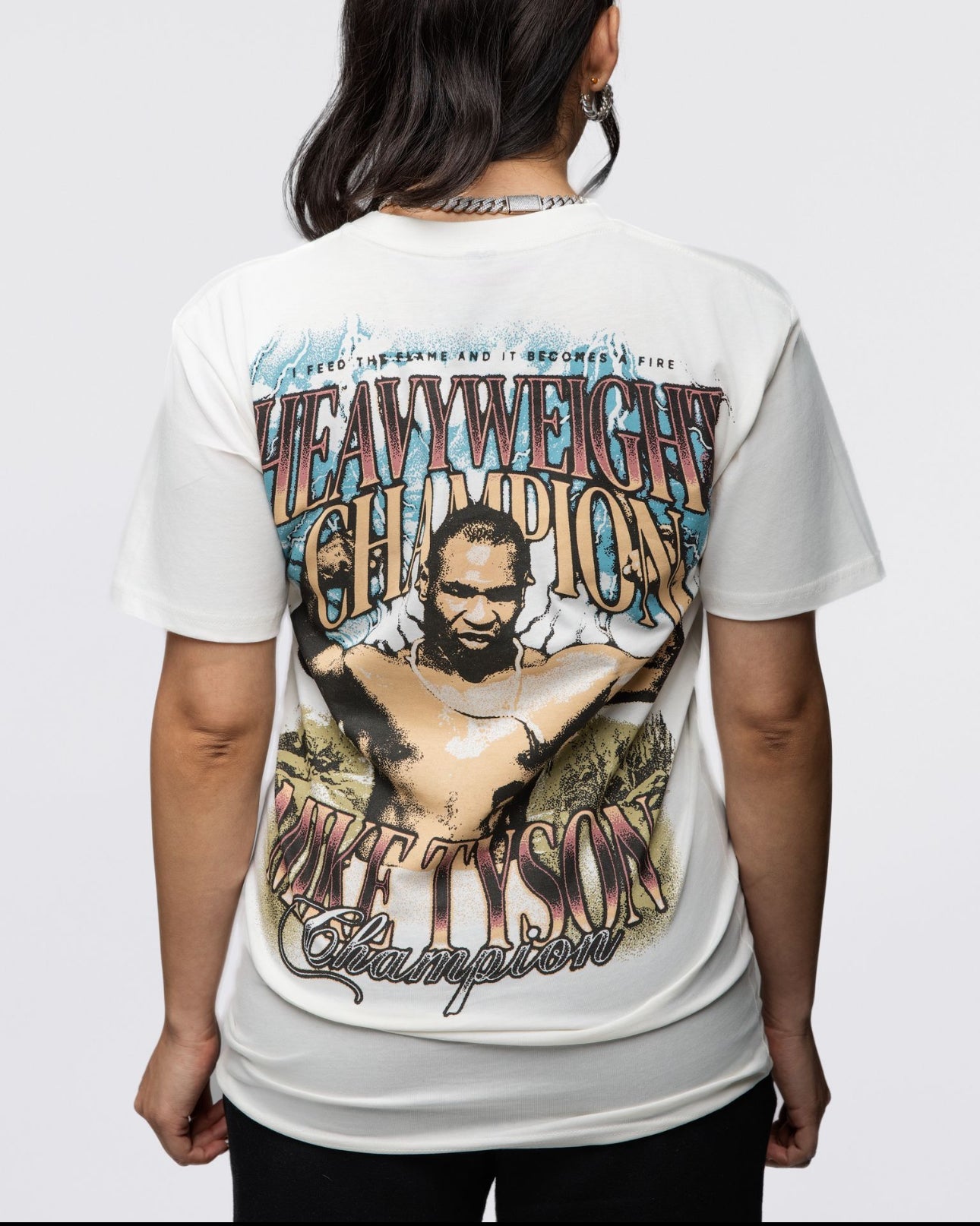 The front of this white shirt features the text "Heavyweight Champion" in a bronze ombre. Smaller text above says "I feed the flame and it becomes a fire." The back features the same text as the front but with an image of Mike with his arms flexed, showing his large biceps. Below, there is additional text that says "Mike Tyson Champion."