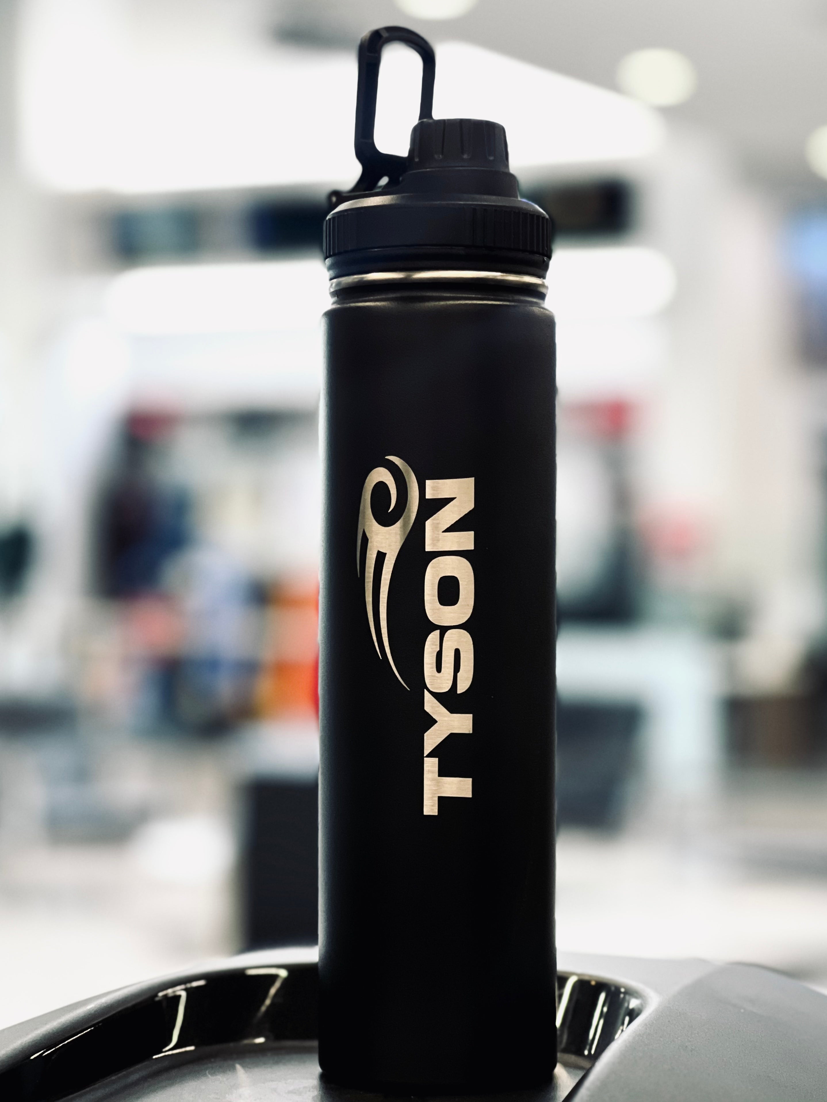 Matte black stainless steel water bottle. Two silver Tyson Pro logos are etched into the center of the bottle running vertically upward on opposite sides. The cap has a loop and the mouthpiece has a screw-top cover.