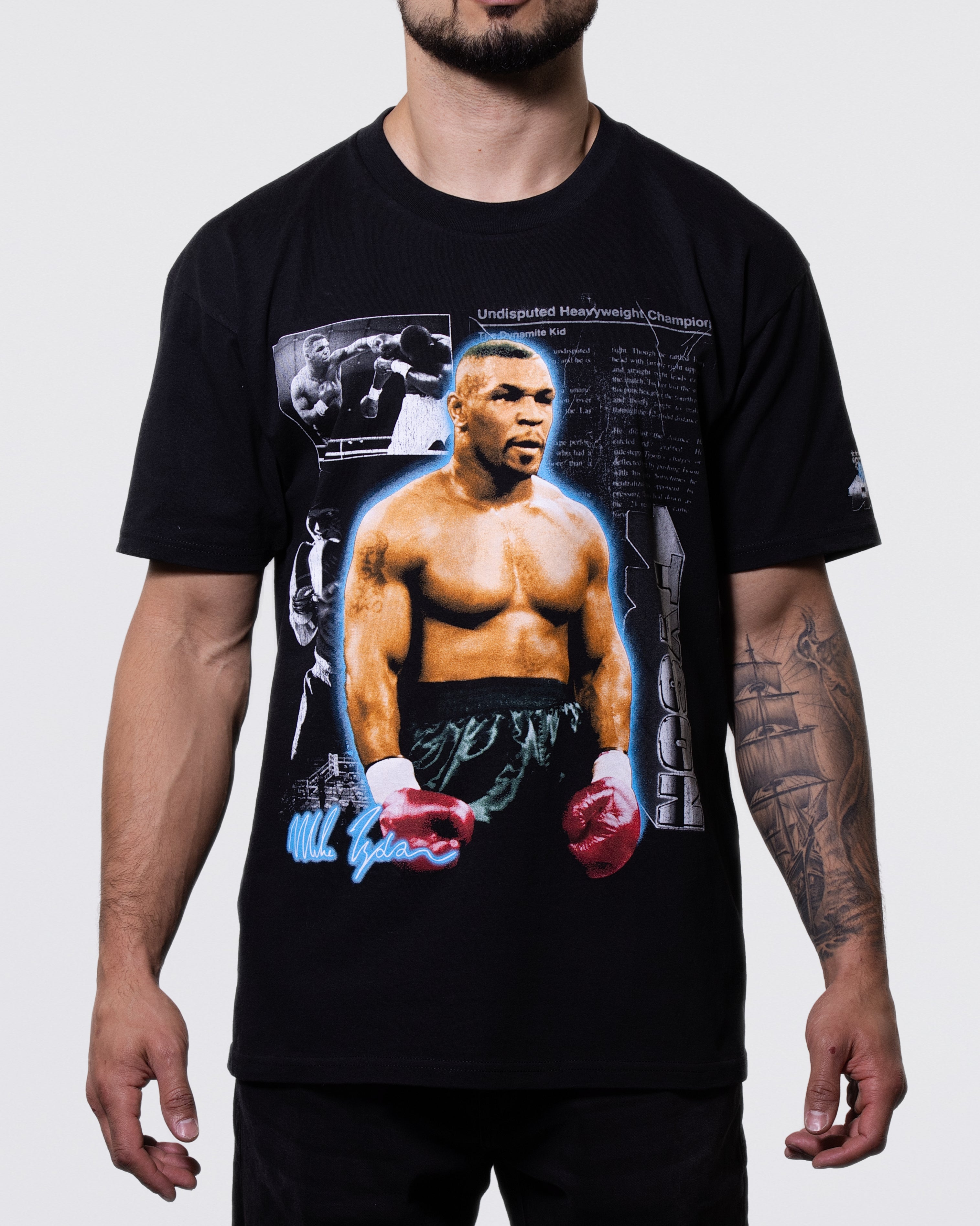 Black T-shirt. There is a large image of Mike in his boxing attire in the center, with smaller images of Mike during fights in the background. There is a faint newspaper clipping also in the background with "Tyson" text on the bottom left and his signature on the bottom right. There is also a small graphic on the left sleeve.